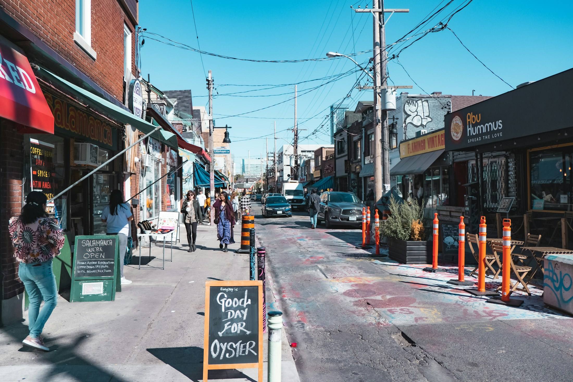 Shoppers on a sunny Kensington Market street lined with colourful storefronts.