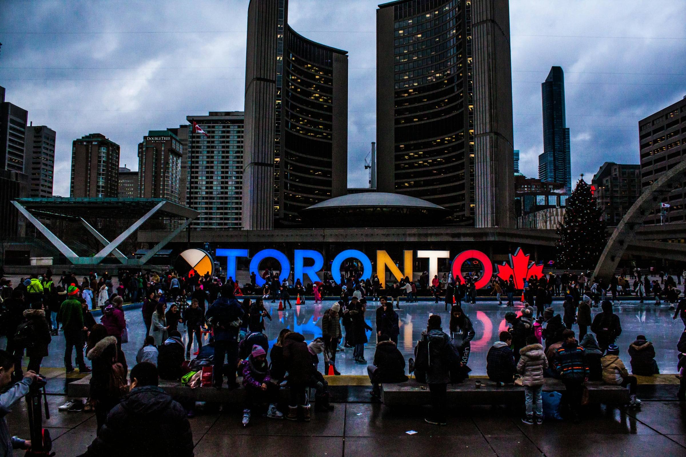 Crowds at Nathan Phillips Square at dusk, with the lit-up TORONTO sign and City Hall's curved towers.